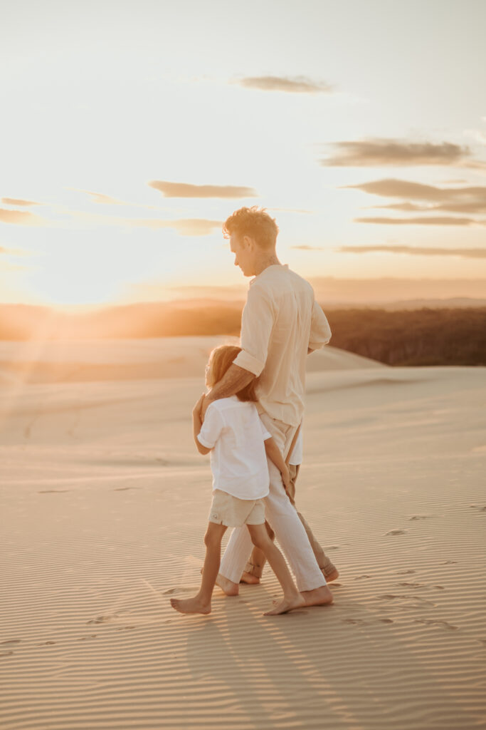 Dad has his arm around his his two young sounds as they stroll together during sunset on the sand dunes.
